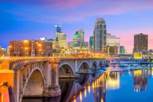 A cityscape view of Minneapolis, Minnesota at dusk, featuring the iconic Stone Arch Bridge spanning the Mississippi River and the city skyline illuminated against a colorful twilight sky.