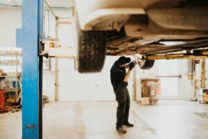 A technician in a black shirt, dark pants, and a cap works under a car lifted on a blue hydraulic lift in a garage. The car's underside and wheels are visible.