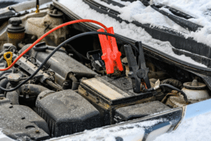 Car battery testing, auto repair in Cokato, MN at Arvu Auto. Image of jumper clamps connected to a vehicle battery in a snow-covered engine bay to support reliable cold-weather starting.