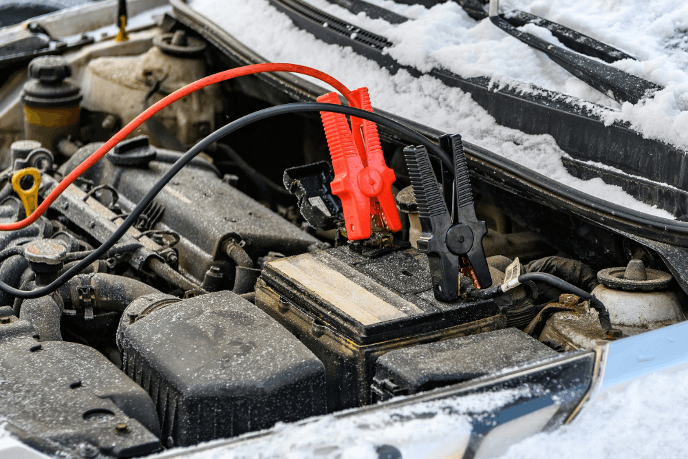 Car battery testing, auto repair in Cokato, MN at Arvu Auto. Image of jumper clamps connected to a vehicle battery in a snow-covered engine bay to support reliable cold-weather starting.