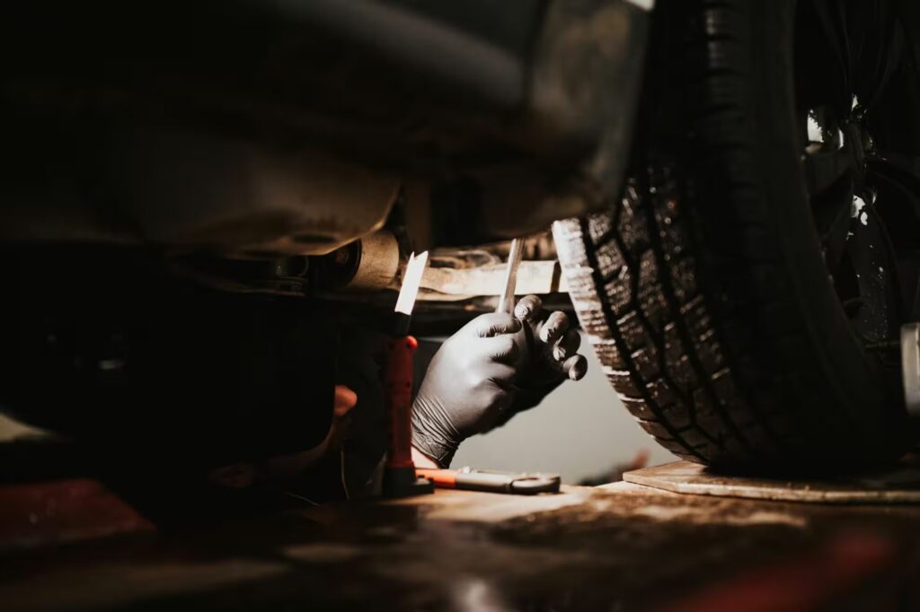 Mechanic wearing black gloves working under a lifted car with a tire in the foreground.