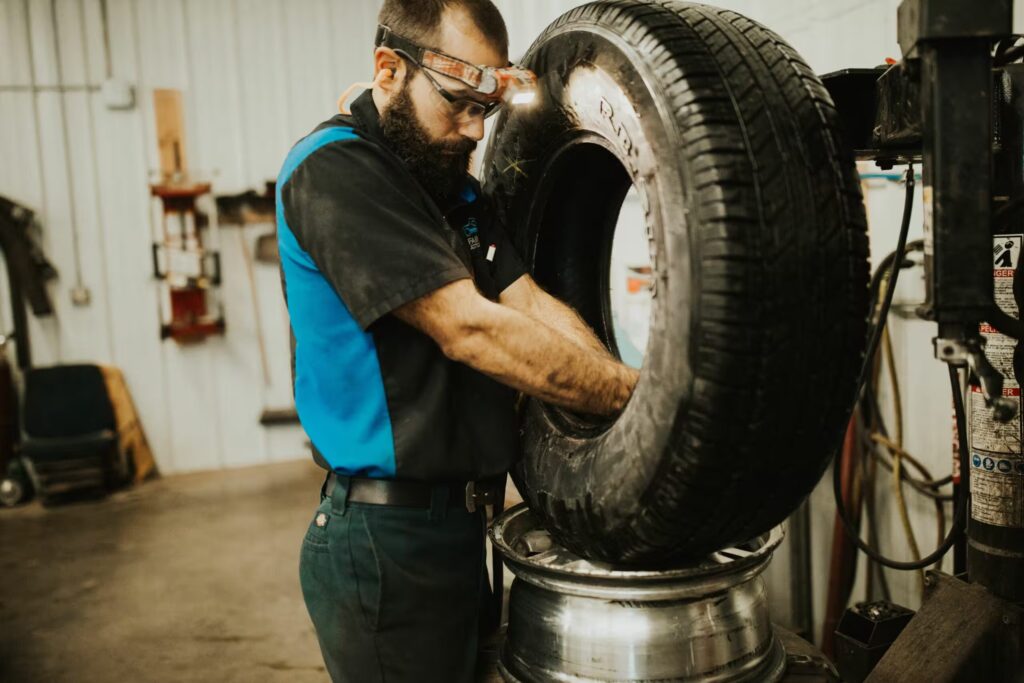 A mechanic wearing safety goggles and gloves installs a tire onto a metal rim using a tire mounting machine in a workshop.