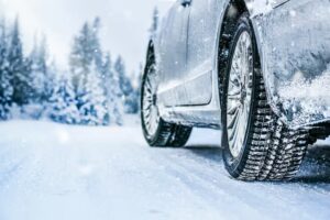 Close-up of a car's winter tires on a snowy road with snow-covered trees in the background.