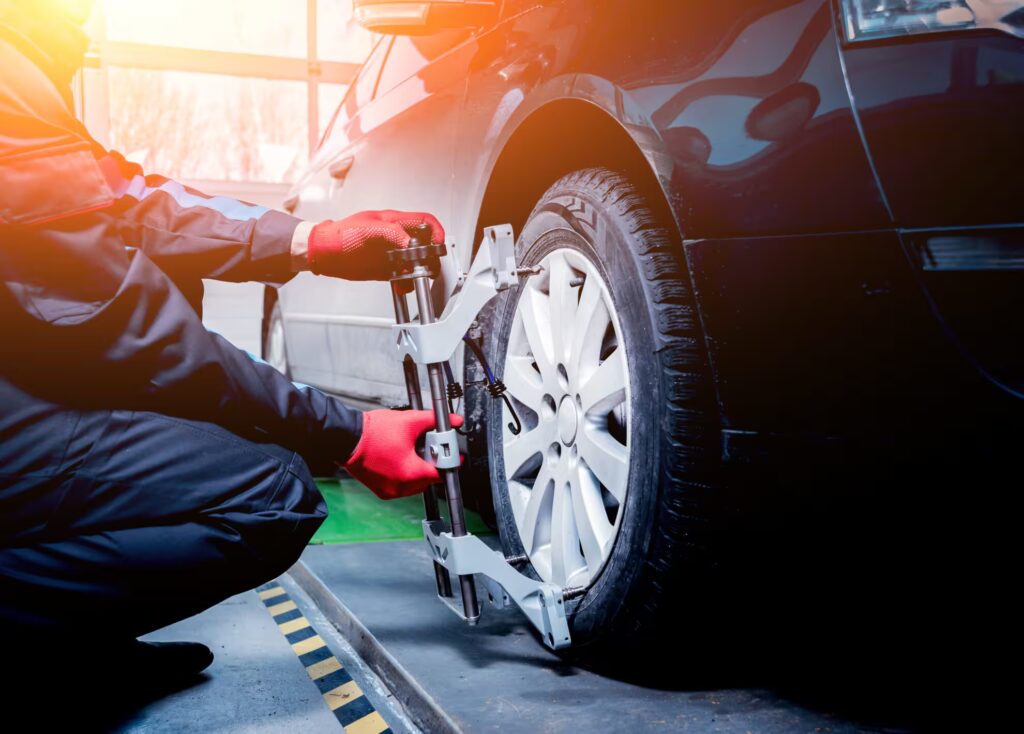 A technician wearing dark work clothes and red gloves is squatting next to a dark-colored car, attaching a wheel alignment tool to the rear wheel.
