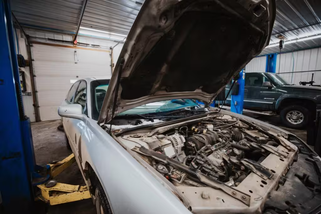 A silver car with its hood open, revealing the engine compartment, is positioned inside an auto repair shop.