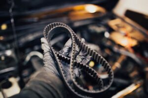 A gloved hand holding a worn timing belt over an open car engine.