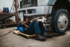 A mechanic lying on a creeper under a dirty truck, working on its undercarriage in a garage.