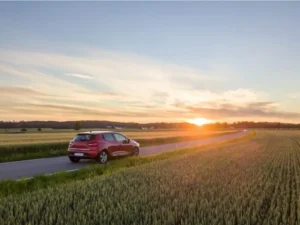 A red hatchback car is driving on an asphalt road through a rural landscape at sunset.