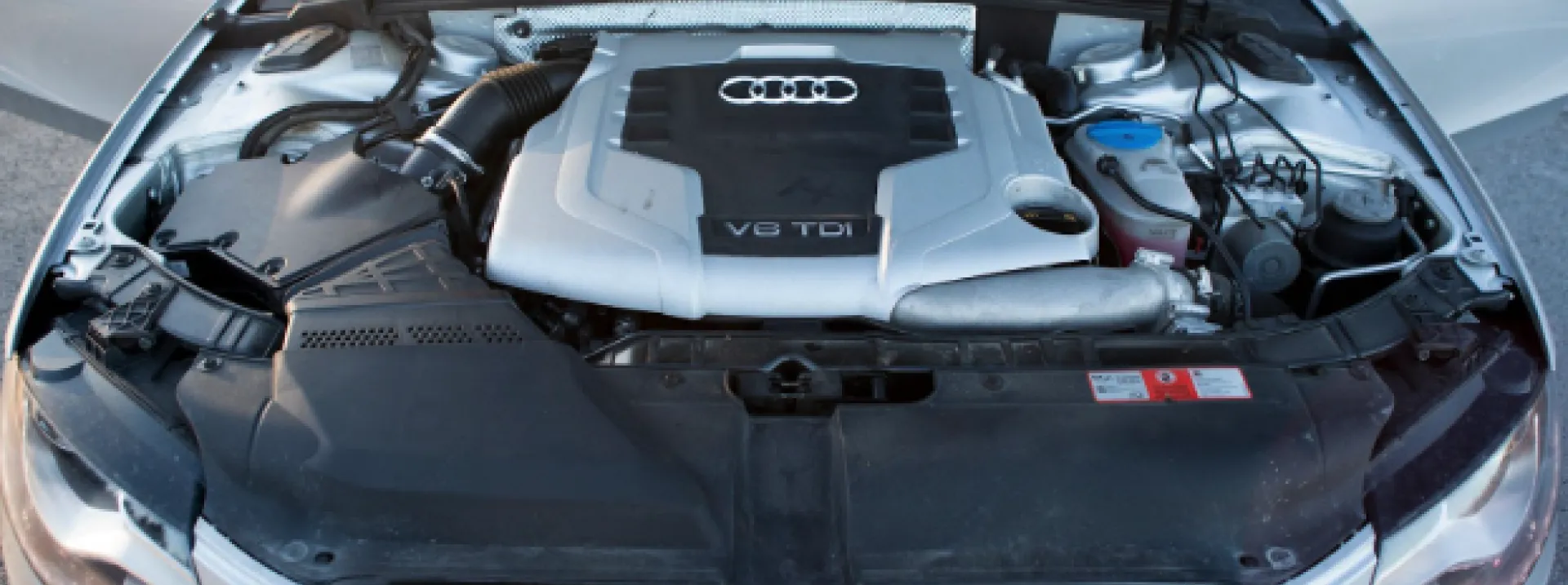 An overhead shot of an Audi A5's engine compartment, revealing the V6 TDI engine and its various components. The engine is centrally located and appears to be in good condition. The surrounding areas are clean and well-organized, with various wires, hoses, and other components visible. The lighting highlights the intricate details of the engine and its surroundings. The image suggests a focus on automotive engineering or maintenance, specifically related to Audi vehicles.