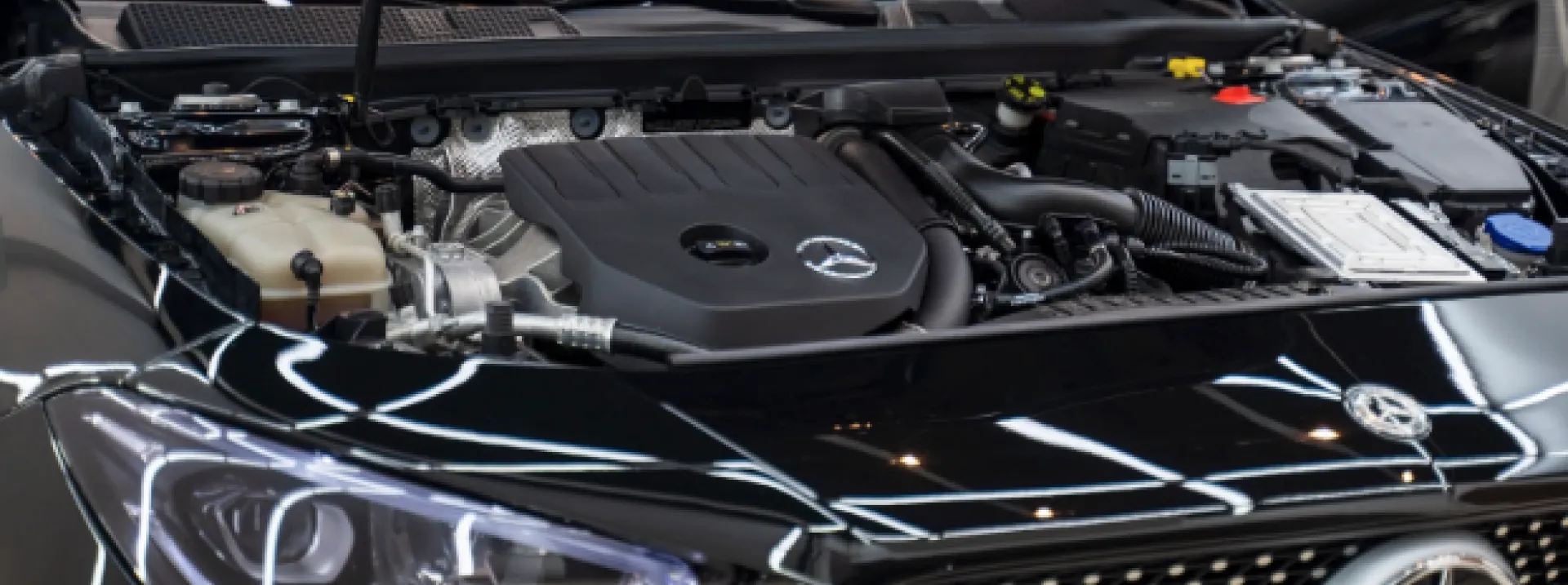 A close-up view of a Mercedes-Benz car engine compartment with the hood open. The engine is centrally located and appears to be in good condition. Various components such as hoses, wires, and fluid reservoirs are visible. The Mercedes-Benz logo is prominently displayed on the engine cover. The lighting highlights the intricate details of the engine and its surroundings. The image suggests a focus on automotive engineering or maintenance, specifically related to Mercedes-Benz vehicles.