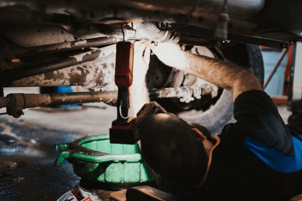 Auto repair near New Germany, MN by Arvu Auto. Image of a mechanic lying under a lifted vehicle, using a light and tools to perform repair work near the exhaust and undercarriage system.